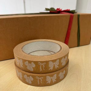 May include: Two rolls of brown washi tape with white bow designs. The tape is wrapped around a cardboard box with a red ribbon.