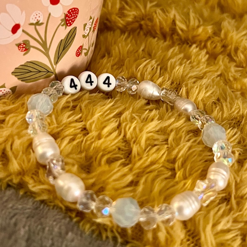 May include: A beaded bracelet with the number "444" spelled out in white letter beads. The bracelet features clear and iridescent beads, along with white pearl-like beads. The background is a fuzzy, golden-yellow texture.