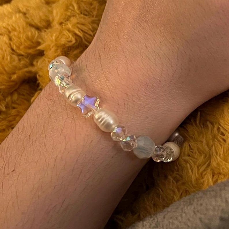 May include: A beaded bracelet featuring white pearls, iridescent crystals, and a star-shaped bead. The bracelet is strung on a clear elastic cord. The bracelet is shown on a wrist.