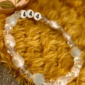 May include: A beaded bracelet featuring white freshwater pearls, clear crystal beads, and light blue beads. The bracelet includes three white letter beads with the number "4" on each. The bracelet is displayed on a yellow, textured surface.
