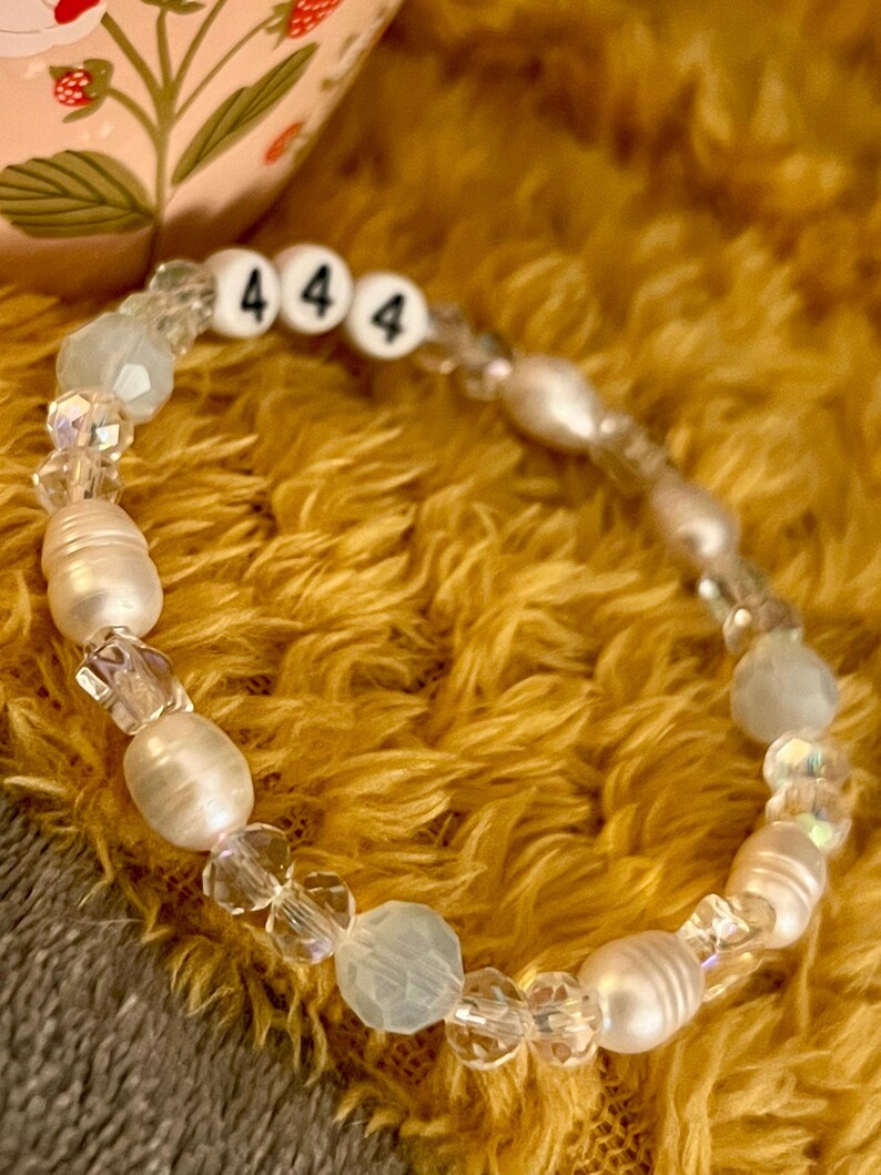 May include: A beaded bracelet featuring white freshwater pearls, clear crystal beads, and light blue beads. The bracelet includes three white letter beads with the number "4" on each. The bracelet is displayed on a yellow, textured surface.