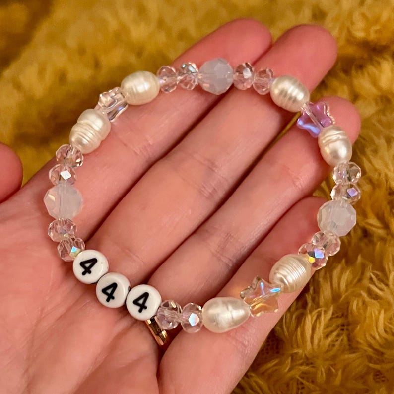May include: A beaded bracelet featuring white freshwater pearls, clear crystal beads, and iridescent accents. The bracelet includes white letter beads spelling out "444". The bracelet is held in a hand.