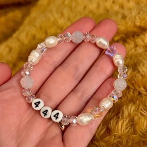 May include: A beaded bracelet featuring white freshwater pearls, clear crystal beads, and iridescent star-shaped beads. The bracelet includes white round beads with the black numbers "444". The bracelet is held in a hand.