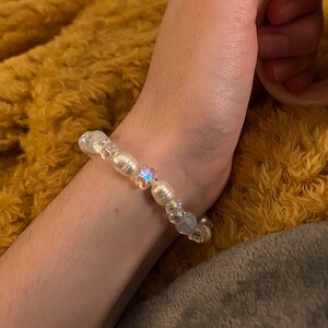 May include: A beaded bracelet featuring iridescent and pearl-like beads. The bracelet includes a star-shaped bead and is worn on a wrist. The background is a fuzzy, golden-yellow blanket.