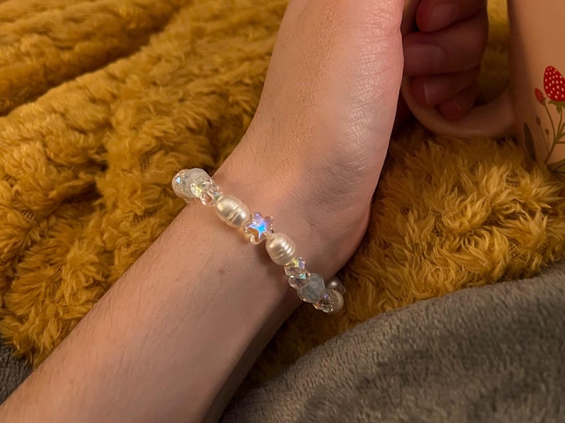 May include: A beaded bracelet featuring iridescent and pearl-like beads. The bracelet includes a star-shaped bead and is worn on a wrist. The background is a fuzzy, golden-yellow blanket.