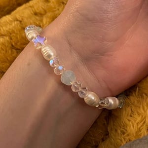 May include: A delicate bracelet featuring a mix of white freshwater pearls, iridescent star-shaped beads, and clear crystal accents. The bracelet is strung on a clear elastic cord, creating a subtle and elegant accessory.