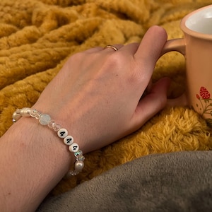 May include: A white beaded bracelet with the numbers "444" on it. The bracelet has clear and iridescent beads. The bracelet is on a person's wrist, with a peach-colored mug with strawberry designs in the background.