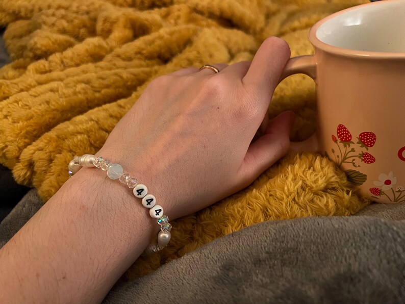 May include: A white beaded bracelet with the numbers "444" on it. The bracelet has clear and iridescent beads. The bracelet is on a person's wrist, with a peach-colored mug with strawberry designs in the background.