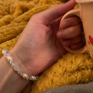 May include: A delicate bracelet with white pearls and iridescent beads. The bracelet is on a wrist, next to a pink mug decorated with red strawberries. The background is a yellow, fluffy blanket.