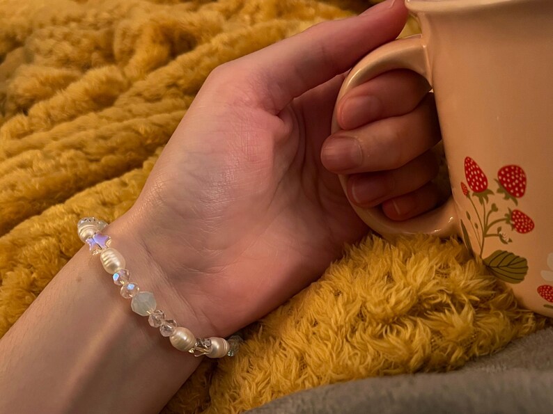 May include: A delicate bracelet with white pearls and iridescent beads. The bracelet is on a wrist, next to a pink mug decorated with red strawberries. The background is a yellow, fluffy blanket.