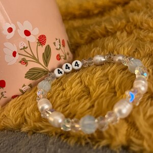 May include: A beaded bracelet with white pearls, clear and blue beads, and white letter beads spelling "444". The bracelet is on a yellow, fuzzy surface. A pink mug with a floral design is in the background.