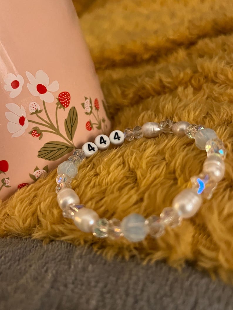 May include: A beaded bracelet with white pearls, clear and blue beads, and white letter beads spelling "444". The bracelet is on a yellow, fuzzy surface. A pink mug with a floral design is in the background.
