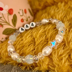 May include: A beaded bracelet featuring white freshwater pearls, iridescent crystals, and star-shaped beads. The bracelet includes beads with the number "444" in black letters. The bracelet is displayed on a yellow, textured surface.