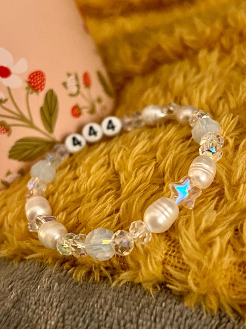 May include: A beaded bracelet featuring white freshwater pearls, iridescent crystals, and star-shaped beads. The bracelet includes beads with the number "444" in black letters. The bracelet is displayed on a yellow, textured surface.