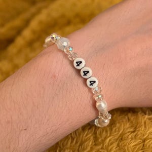 May include: A beaded bracelet with white beads, clear faceted beads, and white letter beads spelling out "444". The bracelet is worn on a wrist against a yellow background.