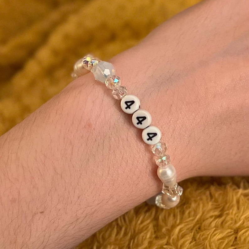 May include: A beaded bracelet with white beads, clear faceted beads, and white letter beads spelling out "444". The bracelet is worn on a wrist against a yellow background.