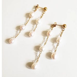 Pode incluir: Um par de brincos de ouro com pérolas brancas. Cada brinco tem uma corrente com quatro pérolas penduradas.