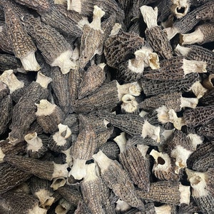 May include: A close-up view of a large pile of dried morel mushrooms. The mushrooms are a dark brown color and have a honeycomb-like texture.