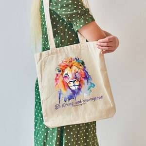Op de afbeelding: Een witte canvas tote bag met een kleurrijk aquarel schilderij van een leeuwenkop en de tekst "Wees sterk en moedig Deuteronomium 31:6".