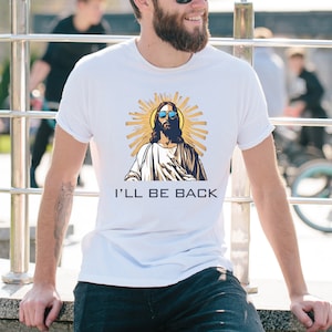 以下が含まれることがあります： サングラスをかけたイエスのグラフィックが描かれた白いTシャツ。金色の光輪が描かれています。「I'LL BE BACK」の文字が黒でプリントされています。柔らかい素材で作られたシャツです。
