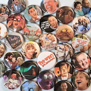 May include: A collection of 25 round buttons featuring various characters and scenes from popular 80s and 90s movies and television shows. The buttons are colorful and feature images of Pee-wee Herman,  'Don't Leave the House Today', 'Chuck's Bike-O-Rama', and more.