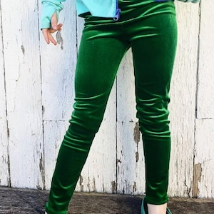 May include: Green velvet leggings paired with teal clogs. The leggings have a soft, shiny texture and are fitted. The clogs are a solid teal color. A light blue jacket with a striped pattern is visible.