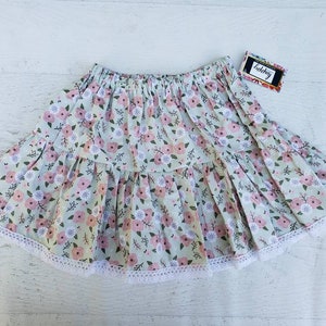 May include: A light green skirt with a floral pattern of pink flowers and white accents. The skirt has a tiered design and is trimmed with white lace along the hemline. The tag says "happy".