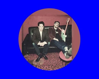May include: Two men seated on a black sofa inside a red-walled room, framed by a blue circle. One man wears a dark blazer and jeans, the other a burgundy jacket, black t-shirt, and jeans, holding a light green guitar. A small red table is nearby.
