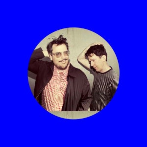 Może przedstawiać: Dwóch mężczyzn w niebieskim okręgu. Jeden mężczyzna nosi okulary, czerwoną i białą koszulę w kratę oraz czarną kurtkę. Drugi mężczyzna nosi czarno-białą koszulę w paski. Obaj mężczyźni dotykają swoich głów.