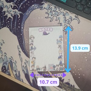 May include: A rectangular notepad, measuring 13.9 cm x 10.7 cm, with a white interior and a border featuring cartoon characters. The top of the notepad has the word "NOTES" and a small purple whale illustration.