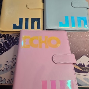 May include: Three leather-like journals in pastel colors: yellow, blue, and pink. Each journal has the word "ECHO" and "JIN" in a holographic, iridescent font. The journals have a snap closure.