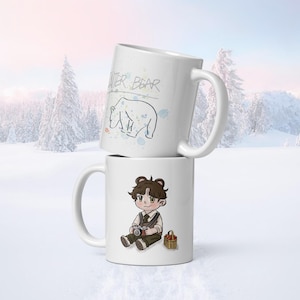 May include: Two white ceramic mugs stacked on top of each other. The top mug has the words "WINTER BEAR" and a bear illustration. The bottom mug features a cartoon character with a camera and a basket of apples.
