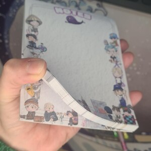 May include: A notepad with a decorative border featuring cartoon characters. The word "NOTED" is at the top, with a purple whale illustration. The notepad is held in a hand, with the pages slightly fanned out.