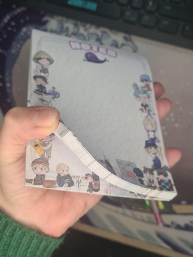 May include: A notepad with a decorative border featuring cartoon characters. The word "NOTED" is at the top, with a purple whale illustration. The notepad is held in a hand, with the pages slightly fanned out.