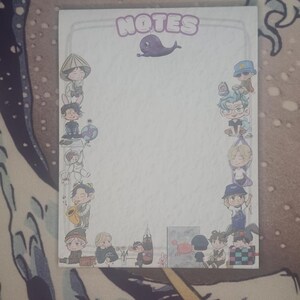 May include: A rectangular notepad featuring the word "NOTES" in purple bubble letters and a cartoon whale illustration. The notepad is bordered by cartoon figures in various outfits. The background is a blue and white patterned surface.