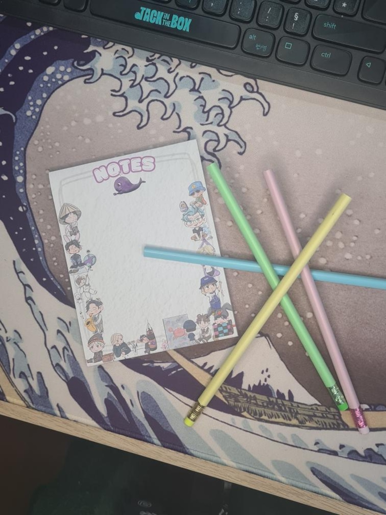 May include: A notepad with the word "NOTES" and a purple whale illustration, surrounded by cartoon characters. Four pastel-coloured pencils are scattered across the notepad. The background is a desk mat with a wave design.