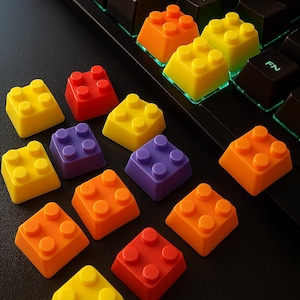 Könnte beinhalten: Ein Satz von 10 bunten Tastenkappen in Form von Lego-Steinen. Die Tastenkappen sind in einem Regenbogenmuster auf einer schwarzen Oberfläche angeordnet. Die Tastenkappen sind aus weichem, strapazierfähigem Kunststoff gefertigt und mit den meisten mechanischen Tastaturen kompatibel.