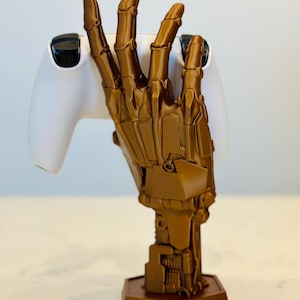 May include: A 3D printed, gold-colored robotic hand holding a white video game controller. The hand is detailed and has a mechanical look. The hand is mounted on a hexagonal base.