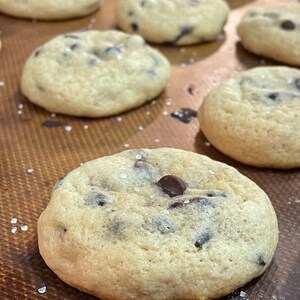 Pu&ograve; includere: Primo piano di un lotto di biscotti al cioccolato appena sfornati con sale marino in cima. I biscotti sono dorati e hanno una consistenza morbida e gommosa.