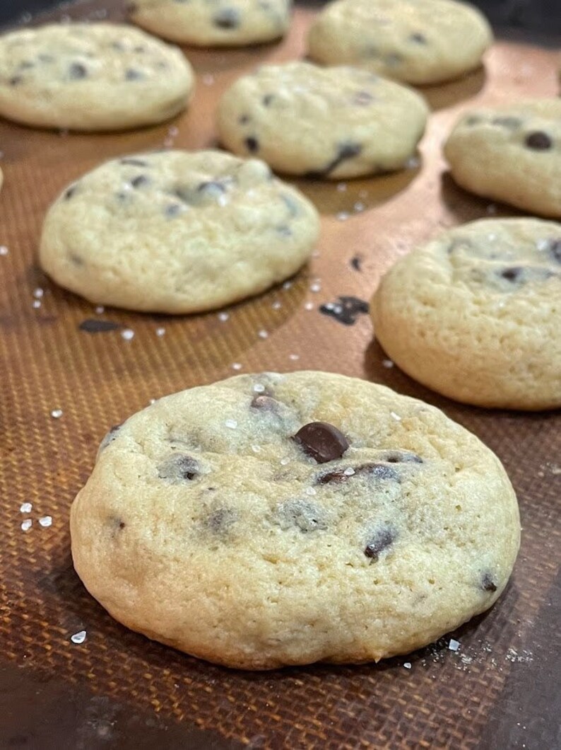 Pu&ograve; includere: Primo piano di un lotto di biscotti al cioccolato appena sfornati con sale marino in cima. I biscotti sono dorati e hanno una consistenza morbida e gommosa.