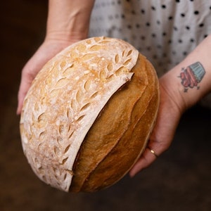 Pu&ograve; includere: Un primo piano di una pagnotta di pane a lievitazione naturale con un motivo decorativo a foglie sulla crosta superiore. Il pane &egrave; dorato e ha una crosta leggermente croccante.