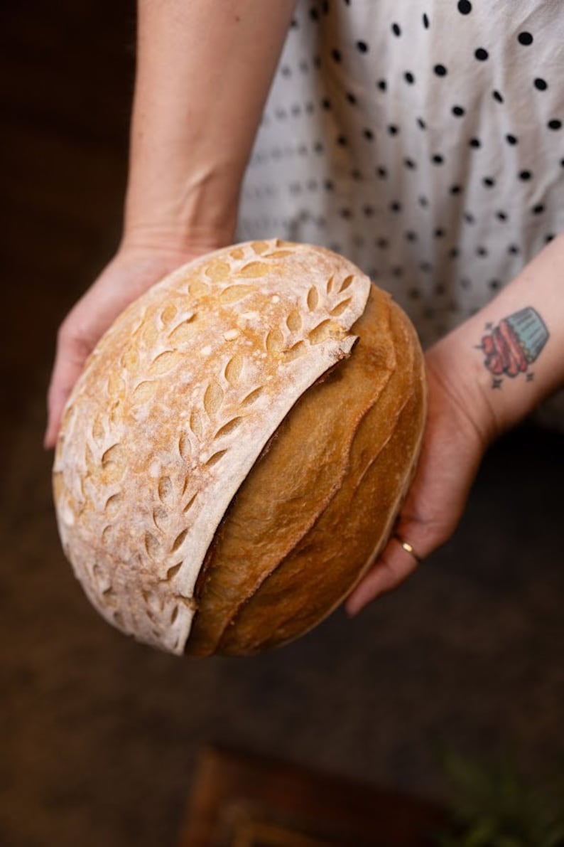 Pu&ograve; includere: Un primo piano di una pagnotta di pane a lievitazione naturale con un motivo decorativo a foglie sulla crosta superiore. Il pane &egrave; dorato e ha una crosta leggermente croccante.
