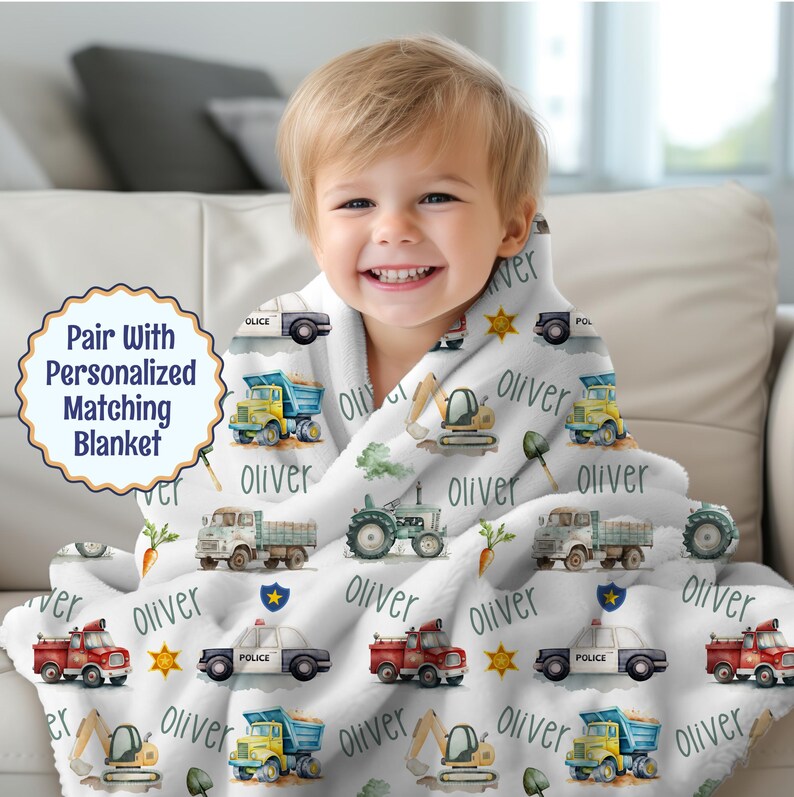 A little boy cuddled up with a blanket with his name and vehicles such as a police car, fire truck, dump truck, farm truck, tractor and excavator.