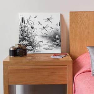 Canvas art on a bedside tabel. The canvas wall art features an elegant black and white line art scene of dragonflies gracefully hovering over a tranquil pond.