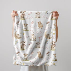 Two hands holding a  personalized custom name white velveteen blanket with whimsical lambs, pink bows, stars, flowers, hearts and butterflies.