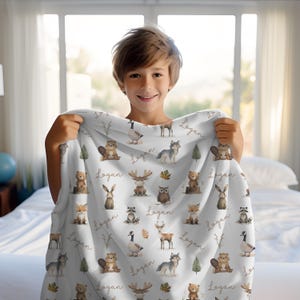 May include: White blanket with woodland animal illustrations, such as bears, foxes, and wolves. The blanket has the name "Logan" printed in a script font. The design includes trees and leaves.