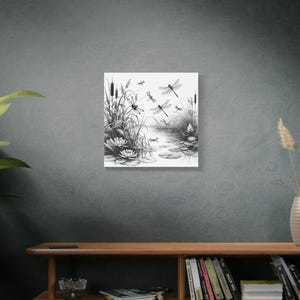 Wall canvas on a grey wall above a low bookcase. Canvas wall art is of elegant black and white line art scene of dragonflies gracefully hovering over a tranquil pond.
