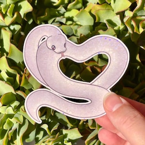 May include: A die-cut sticker featuring a light lavender snake with a subtle holographic glitter finish. The snake is coiled into a heart shape, with its head positioned at the top left and its tail curving to form the bottom point of the heart.