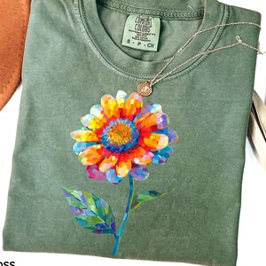 以下が含まれることがあります： モスグリーンの半袖Tシャツで、前面に鮮やかなマルチカラーのモザイク風の花のグラフィックが描かれています。クルーネックで、襟の内側にはComfort Colorsのブランドラベルがあり、サイズSと記載されています。