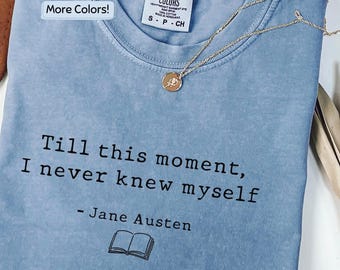 May include: A light blue t-shirt featuring the black printed text, "Till this moment, I never knew myself - Jane Austen" above a small book icon. The shirt has a Comfort Colors brand label at the neck and is paired with a thin gold chain necklace.